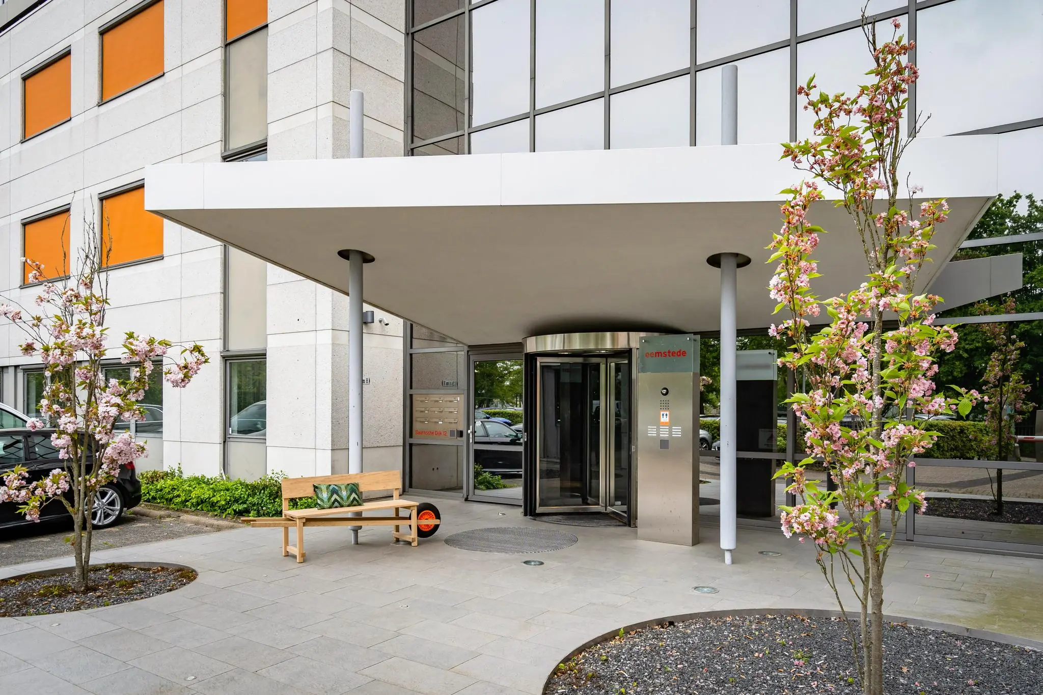 Ingang van het kantoorgebouw aan de Baarnsche Dijk met bloeiende bomen, een bankje en een glazen draaideur.